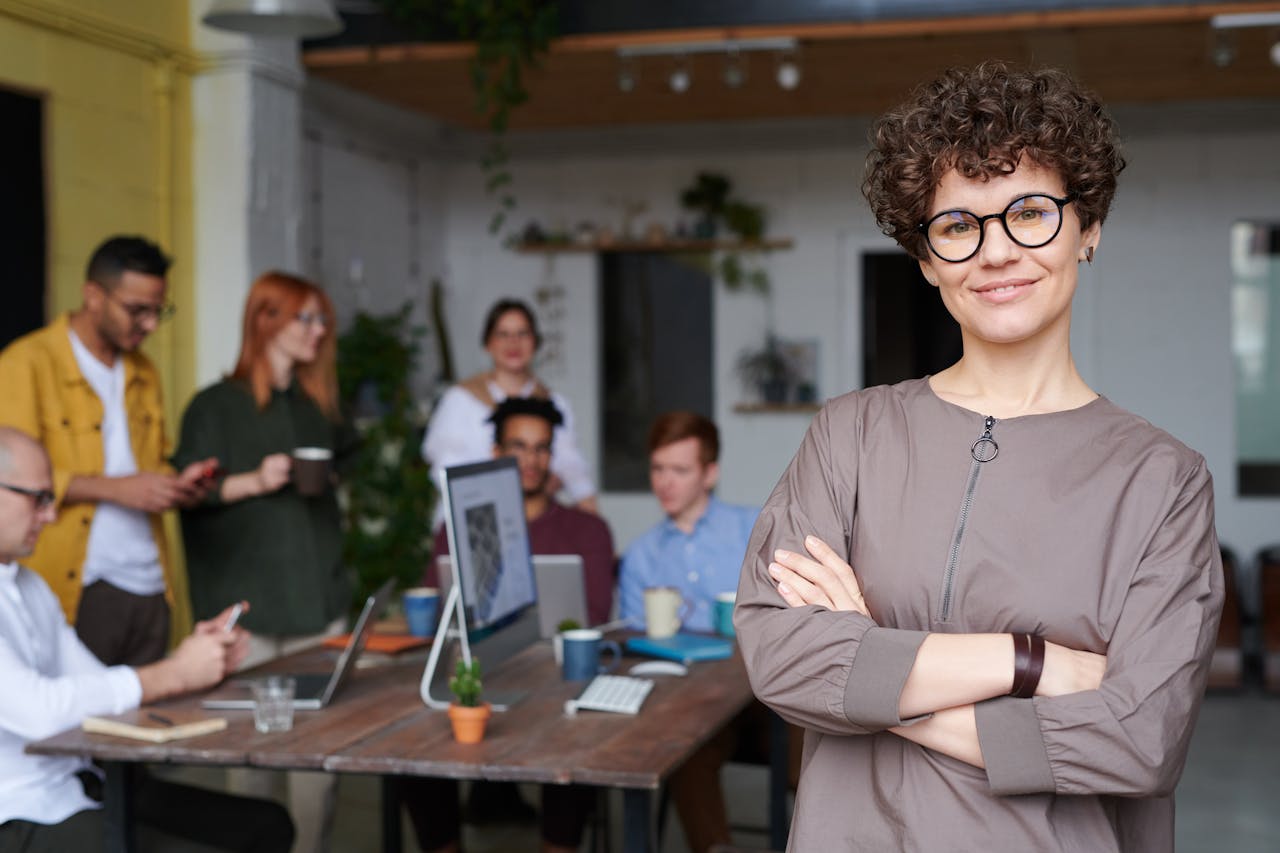 service-05 Smiling businesswoman with curly hair stands confidently in a modern office space with colleagues.