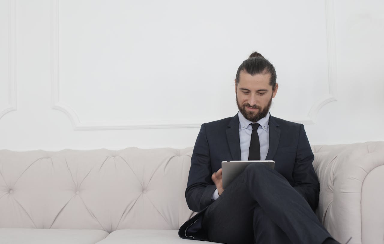 service-04 A businessman in a suit sitting on a sofa using a tablet, smiling.