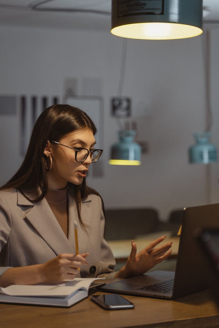 who-we-are A young businesswoman working late at her desk, engaged in an online meeting with a laptop, notebook, and smartphone.